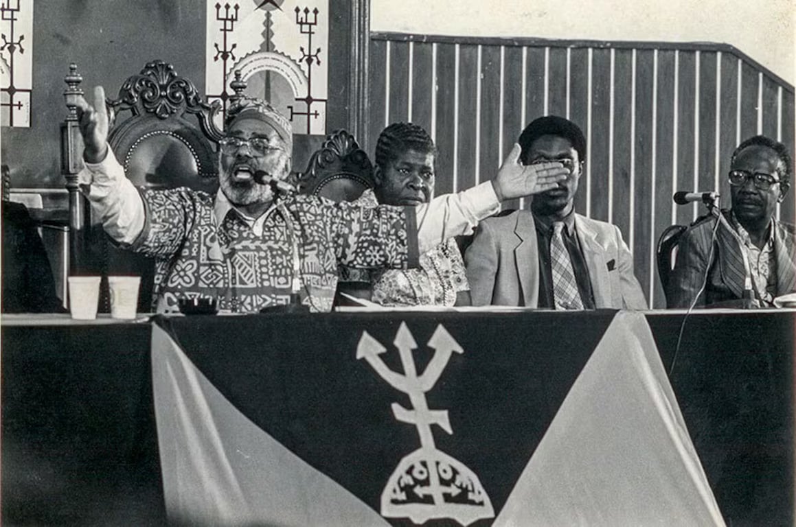 Fotografia em preto e branco do senador Abdias do Nascimento em plenária, discursando com expressão firme e braços estendidos. Ele veste uma camisa estampada com padrões africanos e está sentado ao centro de uma mesa com uma bandeira decorada com símbolo de Exú. Ao fundo, outras lideranças negras acompanham a fala. A imagem transmite força, mobilização e protagonismo político do movimento negro no século XX.