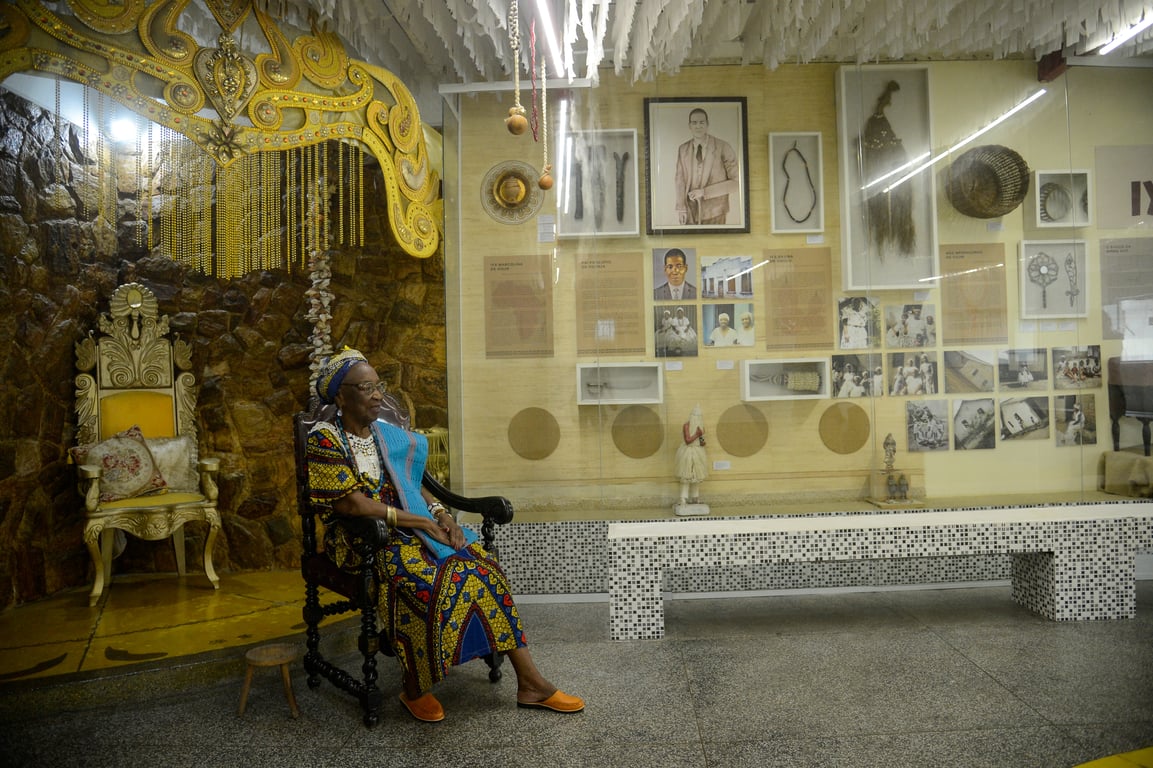 Fotografia colorida tirada no interior do terreiro Ilê Omolu Oxum. No lado esquerdo da imagem, Mãe Meninazinha de Oxum, uma mulher idosa negra, está sentada em uma cadeira de madeira escura, entalhada. Ela veste uma roupa tradicional africana colorida, predominantemente azul, amarelo e vermelho, além de colares brancos e um pano azul sobre o ombro. Usa também um turbante estampado na cabeça e calça sapatos confortáveis de cor laranja. Atrás dela, há uma grande cadeira ornamentada em tom dourado, com detalhes curvos e almofadas decorativas. Acima, pendem franjas douradas e um adorno grande em formato de coroa, também dourado, com desenhos detalhados. A parede ao fundo é revestida com pedras em tonalidade marrom. À direita da foto, há uma longa parede com uma grande vitrine de vidro exibindo fotografias, textos, objetos tradicionais, peças religiosas e instrumentos. Entre as imagens expostas, há retratos antigos, imagens de rituais, artefatos, esculturas e cestos. Alguns objetos pendem do teto, que é coberto por centenas de tiras brancas que criam um efeito texturizado. No chão, um banco comprido revestido de pequenos azulejos pretos e brancos compõe o ambiente. A iluminação é suave, dando destaque à mulher sentada e ao conjunto histórico-cultural ao redor.
