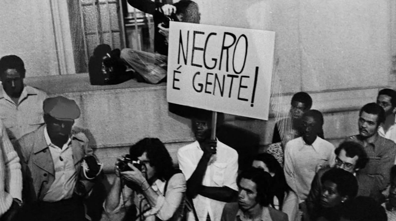 Fotografia em preto e branco registra um protesto do Movimento Negro Unificado (MNU), realizado em 1978. No centro da imagem, um homem negro segura um cartaz com os dizeres 'NEGRO É GENTE!', em letras maiúsculas e firmes. Ele está rodeado por uma multidão atenta, composta por homens e mulheres de diferentes origens, enquanto um fotógrafo registra o momento. Ao fundo, pessoas se apoiam em uma mureta próxima a um prédio de fachada institucional. A cena expressa a força e a urgência da luta antirracista em plena ditadura militar, marco da fundação do MNU — a maior entidade negra da América Latina, surgida em resposta à repressão e à violência policial contra a população negra no Brasil.