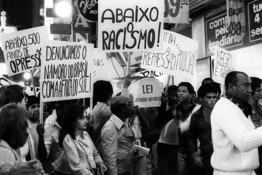 Fotografia em preto e branco do Arquivo Público que registra uma manifestação do movimento negro durante a ditadura militar no Brasil. A cena é tomada por uma multidão de pessoas majoritariamente negras, reunidas em uma rua, segurando cartazes com dizeres de protesto. Algumas placas visíveis dizem: 'Abaixo racismo!', 'Denunciamos o namoro do Brasil com a África do Sul!', 'Abaixo 500 anos de opressão!', 'Negro é gente!', 'Lei Afonso Arinos', entre outras. As expressões dos manifestantes são firmes, com gestos de denúncia, em um momento em que manifestações políticas eram fortemente reprimidas. A imagem evidencia a luta antirracista mesmo sob o regime autoritário, marcada por coragem e articulação coletiva.
