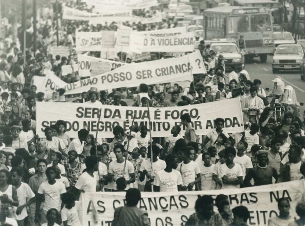 A fotografia em preto e branco mostra uma multidão compacta de pessoas, sendo crianças, outros adolescentes e adultos, caminhando em uma rua, com faixas sendo carregadas na frente. A composição é centrada, com as pessoas ocupando a maior parte do quadro. As ações são da Manifestação pelos direitos da infância e da juventude nos anos 1980, onde as pessoas marcham e carregam faixas com mensagens.