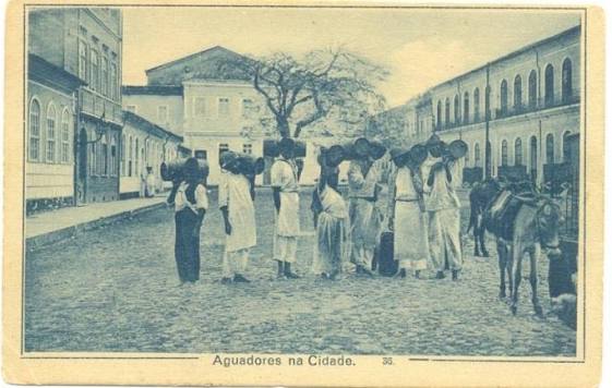 Fotografia intitulada Aguadores na Cidade, pertencente ao acervo da Fundação Gregório de Matos. A imagem mostra um grupo de homens e mulheres negros reunidos em fila junto a um chafariz, aguardando para buscar água. A maioria carrega grandes barris apoiados sobre a cabeça, enquanto outros seguram recipientes ao lado do corpo. As pessoas vestem roupas claras e compridas, típicas da época. À direita, aparece um animal de carga parado próximo ao grupo. A cena acontece em uma rua de paralelepípedos, cercada por casarões de dois andares, com um grande chafariz e uma árvore ao fundo. Na parte inferior da fotografia, lê-se a legenda “Aguadores na Cidade”. O registro retrata o trabalho dos aguadores, responsáveis pelo transporte e abastecimento de água nas cidades brasileiras durante o século XIX.