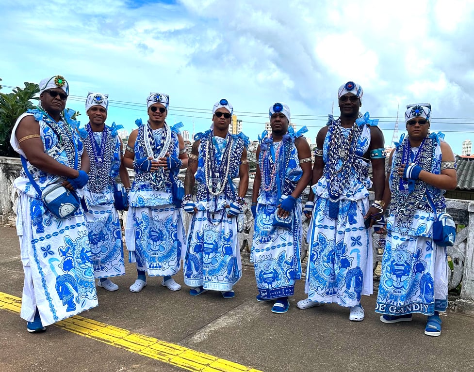 Fotografia de associados do Afoxés Filhos de Gandhy durante o Carnaval de 2026. A imagem mostra sete homens negros alinhados lado a lado, posando para a câmera em uma área aberta. Todos vestem as tradicionais roupas brancas e azuis, com saias estampadas, turbantes brancos adornados com detalhes azuis, colares de contas, faixas, pulseiras e bolsas atravessadas no corpo. Alguns usam óculos escuros e calçados nas cores branca e azul. As vestimentas são ricamente ornamentadas com símbolos e estampas características dos Filhos de Gandhy. Ao fundo, aparecem o céu com muitas nuvens, construções urbanas e parte da paisagem de Salvador. A fotografia registra integrantes do tradicional Afoxés Filhos de Gandhy, um dos principais símbolos do Carnaval e da cultura afro-brasileira na Bahia.