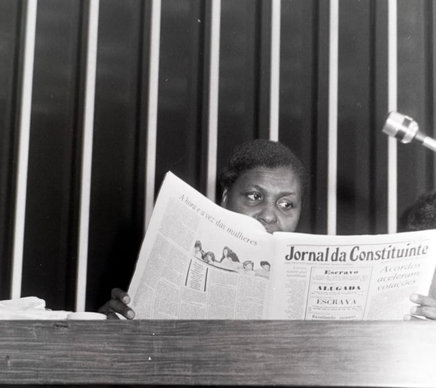 Foto da deputada federal Benedita da Silva, uma mulher negra de meia-idade, segurando o jornal aberto em suas mãos. Ela está situada em frente a um fundo com linhas verticais claras e escuras, que indicam um painel de madeira ou algum tipo de estrutura. A presença de um microfone no canto direito sugere um contexto de discurso futuro.