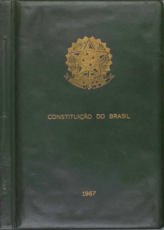 Fotografia da capa da Constituição do Brasil de 1967. O fundo é em couro verde-escuro, com textura levemente enrugada. No centro, em dourado, está o brasão da República Federativa do Brasil, composto por uma estrela de cinco pontas ladeada por ramos de café e fumo, com a inscrição 'República Federativa do Brasil' em uma faixa inferior. Logo abaixo, em letras douradas, lê-se 'CONSTITUIÇÃO DO BRASIL'. Na parte inferior central, também em dourado, está o ano '1967'. A encadernação é lateral, com dois pequenos rebites visíveis à esquerda.