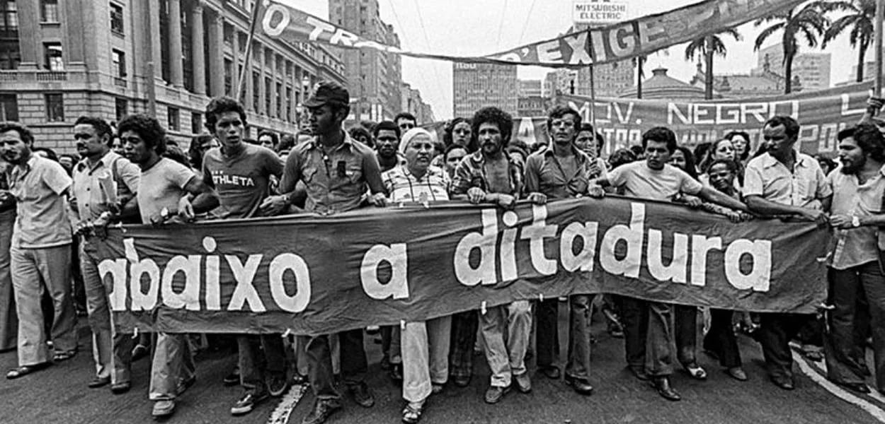 Fotografia em preto e branco de uma manifestação popular no período da ditadura militar no Brasil. Um grupo de homens e mulheres caminha lado a lado segurando uma grande faixa estendida na horizontal, onde se lê em letras brancas: abaixo a ditadura. Ao fundo, outras faixas e cartazes também aparecem erguidos, com dizeres parcialmente visíveis como MOV. NEGRO e EXIGÊ…. A cena acontece em uma avenida de cidade grande, com prédios altos e palmeiras ao fundo. Os manifestantes ocupam toda a largura da rua, com expressões de seriedade e resistência.