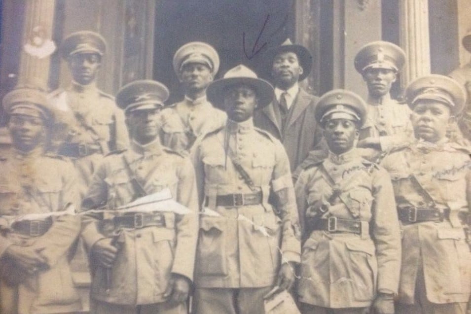 A imagem mostra um grupo de homens negros posando para uma fotografia formal em frente a um edifício, provavelmente institucional. A maioria deles veste uniformes militares completos, jaquetas, cintos, quepes ou chapéus, típicos do início do século XX. Os uniformes incluem insígnias e botões alinhados, sugerindo organização e hierarquia dentro do grupo. Na fileira da frente, vários soldados encaram diretamente a câmera, alguns com expressão séria, outros com semblante mais neutro. Ao centro, um homem usa um chapéu de aba larga, diferente dos quepes dos demais, destacando-se visualmente. Logo atrás dele, há um homem vestido de terno escuro e chapéu, que não usa uniforme; acima de sua cabeça há uma seta desenhada à mão, indicando que ele é uma figura específica ou importante para quem marcou a foto. O fundo mostra colunas e portas, possivelmente de um prédio público ou quartel. A fotografia apresenta sinais de desgaste, arranhões, rasgos e descoloração, evidenciando sua antiguidade. Trata-se de um registro histórico de integrantes da Legião Negra, unidade formada por soldados negros durante a Revolução Constitucionalista de 1932.