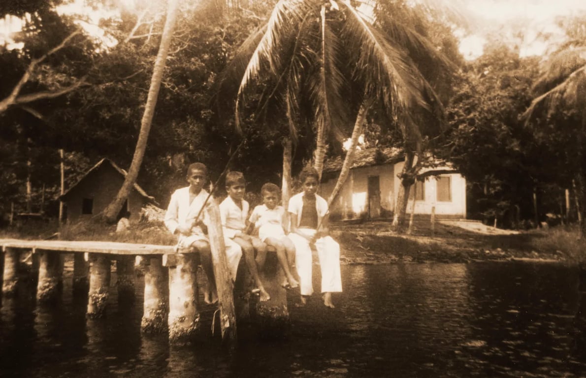 Fotografia em tom sépia, com aparência antiga, mostra quatro crianças negras sentadas na beira de um trapiche de madeira sobre um rio, durante uma pescaria. Três delas vestem roupas claras e uma segura um varal de pesca. As crianças estão descalças e com os pés pendurados sobre a água. Ao fundo, há casas simples com telhado de palha e paredes de taipa, cercadas por vegetação tropical densa e coqueiros altos. A cena transmite tranquilidade e remete à vida cotidiana em comunidades ribeirinhas ou quilombolas, possivelmente no interior do Brasil, em um tempo passado. A luz suave e o enquadramento reforçam o clima de memória afetiva e ancestralidade.