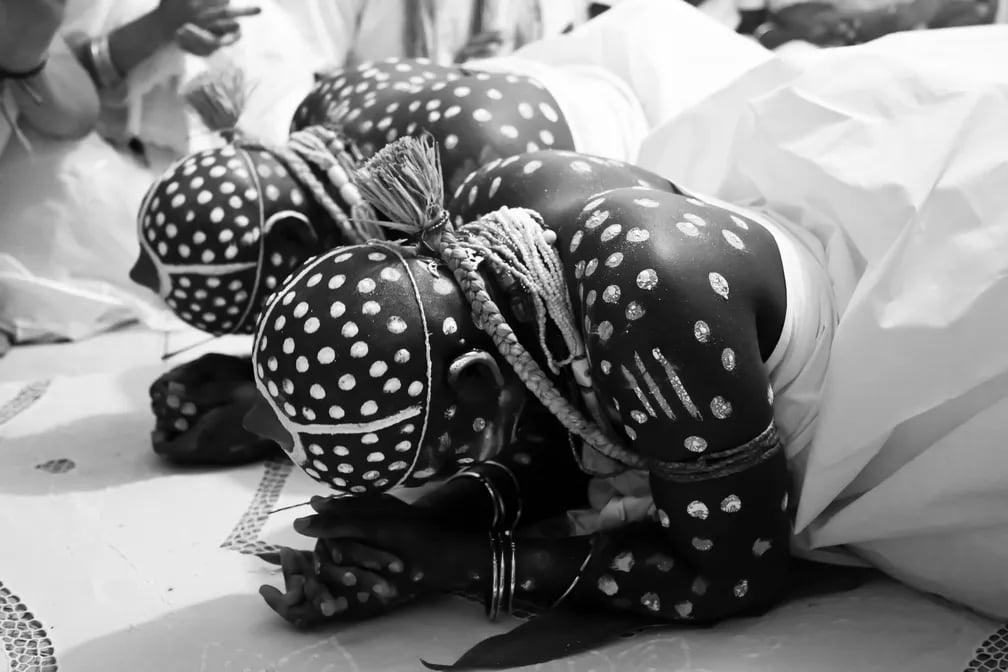 Fotografia em preto e branco intitulada Candomblé da Bahia, de Adeloyá Magnoni, pertencente ao arquivo pessoal da fotógrafa. A imagem mostra duas pessoas negras deitadas de bruços sobre o chão durante um ritual religioso. Elas vestem roupas brancas e têm a cabeça, os braços e parte do corpo pintados com pontos e linhas brancas. Ambas utilizam fios de contas e pulseiras. As mãos permanecem apoiadas no chão e os rostos voltados para baixo, em posição de reverência. Ao redor, aparecem parcialmente outras pessoas vestidas de branco, acompanhando a cerimônia. A fotografia registra um momento de ritual do candomblé, religião de matriz africana presente na Bahia.