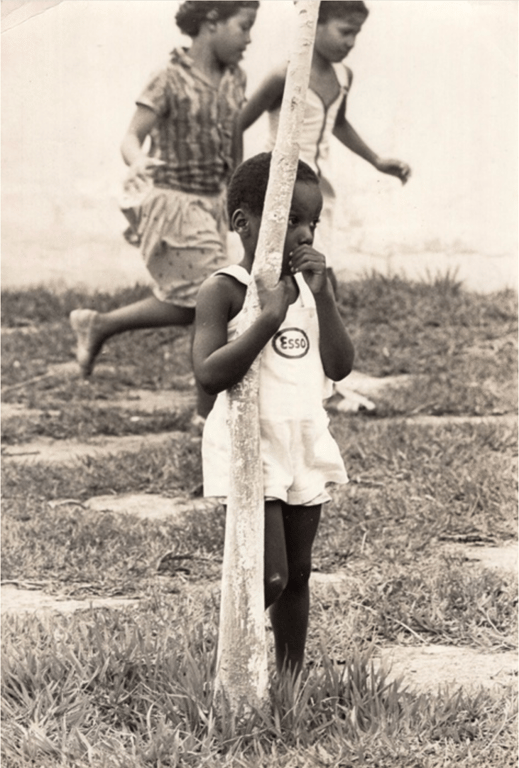 Fotografia em preto e branco de 1979. Em primeiro plano, uma criança negra, vestida com um macacão branco com o logotipo 'Esso' no peito, está encostada em um tronco de árvore pintado de branco, segurando-o com os braços e levando a mão à boca, com expressão pensativa ou tímida. A criança está descalça, em pé sobre a grama. Ao fundo, outras duas crianças, também negras, correm e brincam, desfocadas em movimento. Todas estão em um ambiente externo, com gramado e piso de cimento. A imagem foi registrada durante uma festa de Natal promovida pela FUNABEM (Fundação Nacional do Bem-Estar do Menor), no Rio de Janeiro, e tem o carimbo do 'Jornal do Brasil' no verso.
