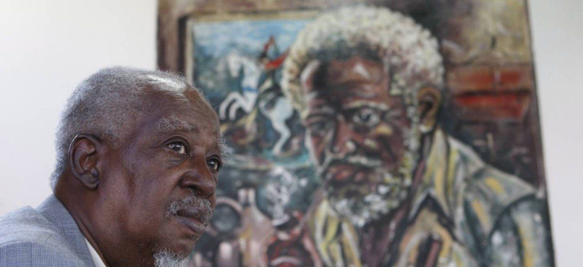 Fotografia colorida de Carlos Alberto Caó, um homem negro idoso, de cabelos grisalhos e expressão serena, usando paletó claro. Ele está em primeiro plano, levemente de perfil, olhando para a esquerda. Ao fundo, desfocada, há uma pintura que retrata um homem negro com traços semelhantes: cabelo branco, barba espessa e expressão introspectiva. A obra ao fundo traz tons escuros e terrosos, com elementos que remetem à cultura afro-brasileira. A imagem transmite um forte senso de ancestralidade, memória e identidade. Foto: Agência O Globo / Eduardo Naddar.