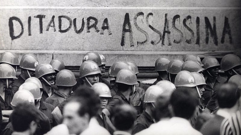 Fotografia em preto e branco registra uma cena de protesto durante o período da ditadura militar no Brasil. No centro da imagem, um grupo de soldados usando capacetes e fardas escuras forma uma barreira diante de uma parede de pedra. Na parede, em letras grandes e marcantes, está pichada a frase "DITADURA ASSASSINA". Em primeiro plano, há manifestantes e civis parcialmente desfocados, observando ou interagindo com os militares. A imagem transmite tensão e resistência, sendo um forte símbolo da repressão estatal e da denúncia popular contra a violência do regime militar.