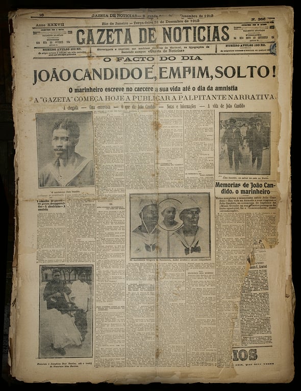 Fotografia de uma página do jornal Gazeta de Notícias, edição de 31 de dezembro de 1912. A diagramação ocupa toda a página com colunas de texto e várias imagens em preto e branco. No topo, em letras garrafais, está o título da matéria principal: 'João Candido é, enfim, solto!', com o subtítulo logo abaixo informando que o marinheiro escreve no cárcere sua vida até o dia da anistia. Abaixo do título há três fotografias em destaque: à esquerda, um retrato de João Cândido com olhar sério; ao centro, uma imagem com três marinheiros fardados; à direita, João Cândido marchando acompanhado por outros homens. Também aparecem imagens menores, como a de duas mulheres. O visual do jornal está amarelado e gasto, com bordas rasgadas e marcas do tempo, evidenciando sua antiguidade. A página transmite a importância histórica do momento, celebrando a libertação de João Cândido após a Revolta da Chibata.
