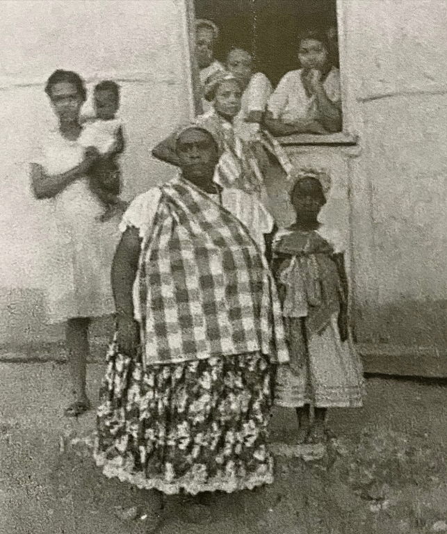 Fotografia em preto e branco mostra um grupo de pessoas negras diante de uma casa simples. Em primeiro plano, ao centro, uma mulher vestida com saia longa florida e pano xadrez sobre os ombros encara a câmera com expressão séria; ao seu lado, uma menina com vestido tradicional. À esquerda, uma mulher segura uma criança no colo. Ao fundo, na janela da casa, outras mulheres observam a cena, algumas vestindo roupas de baiana e com turbantes. A imagem transmite força, ancestralidade e cotidiano, possivelmente registrada em uma comunidade afro-brasileira na primeira metade do século XX.