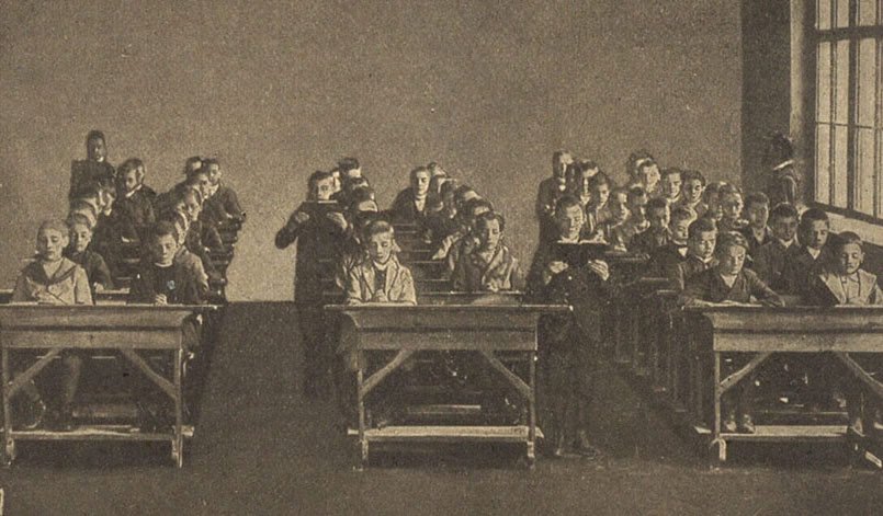 Fotografia em sépia mostrando uma sala de aula antiga, com dezenas de meninos brancos, todos usando roupas escuras formais, sentados em carteiras de madeira enfileiradas. As carteiras têm estrutura robusta e estão organizadas em três colunas. Ao fundo, duas figuras adultas, possivelmente professores ou monitores, observam os alunos. A sala é ampla, com piso escuro e paredes lisas e claras. À direita, uma grande janela deixa entrar luz natural, iluminando parcialmente os rostos dos estudantes. A composição da imagem reforça a rigidez e a formalidade da educação primária voltada às crianças brancas nas instituições públicas da Corte durante o Império, evidenciando a exclusão de crianças negras do sistema educacional da época.