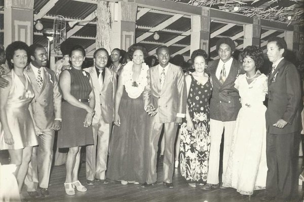 Fotografia em preto e branco do Baile de Cem Anos da entidade Floresta Aurora, realizado em 1972. Em um salão com teto alto e colunas decoradas, aparece um grupo de onze pessoas negras posando lado a lado, todas vestidas de forma elegante e sorrindo para a câmera. As mulheres usam vestidos longos ou curtos e penteados bem arrumados. Os homens vestem ternos bem alinhados, com gravatas e lapelas decoradas. O ambiente está iluminado, com enfeites no teto e pessoas ao fundo, sugerindo um momento festivo. A imagem revela a importância do clube como espaço de celebração, pertencimento e resistência da comunidade negra no Brasil.