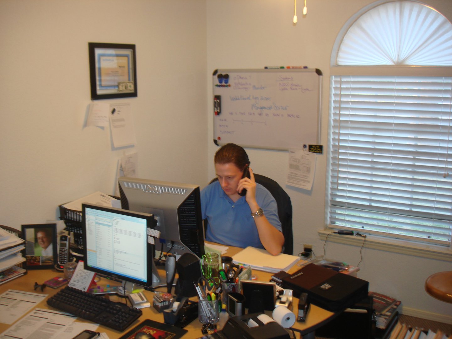 Person working on the phone at a desk in an early office, surrounded by computer monitors, a whiteboard, and office equipment.