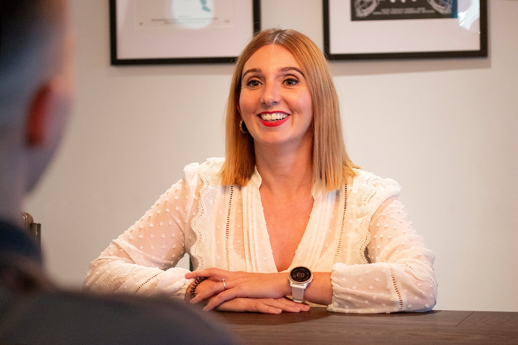 A woman in a white blouse smiling at a blurred figure opposite, with her hands folded over the other