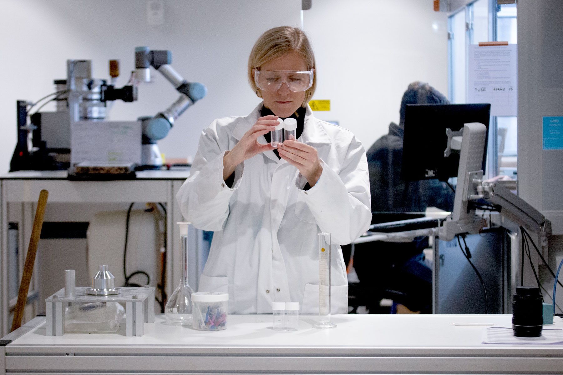 A woman in a laboratory wearing safety goggles and a lab coat inspecting two clear vials