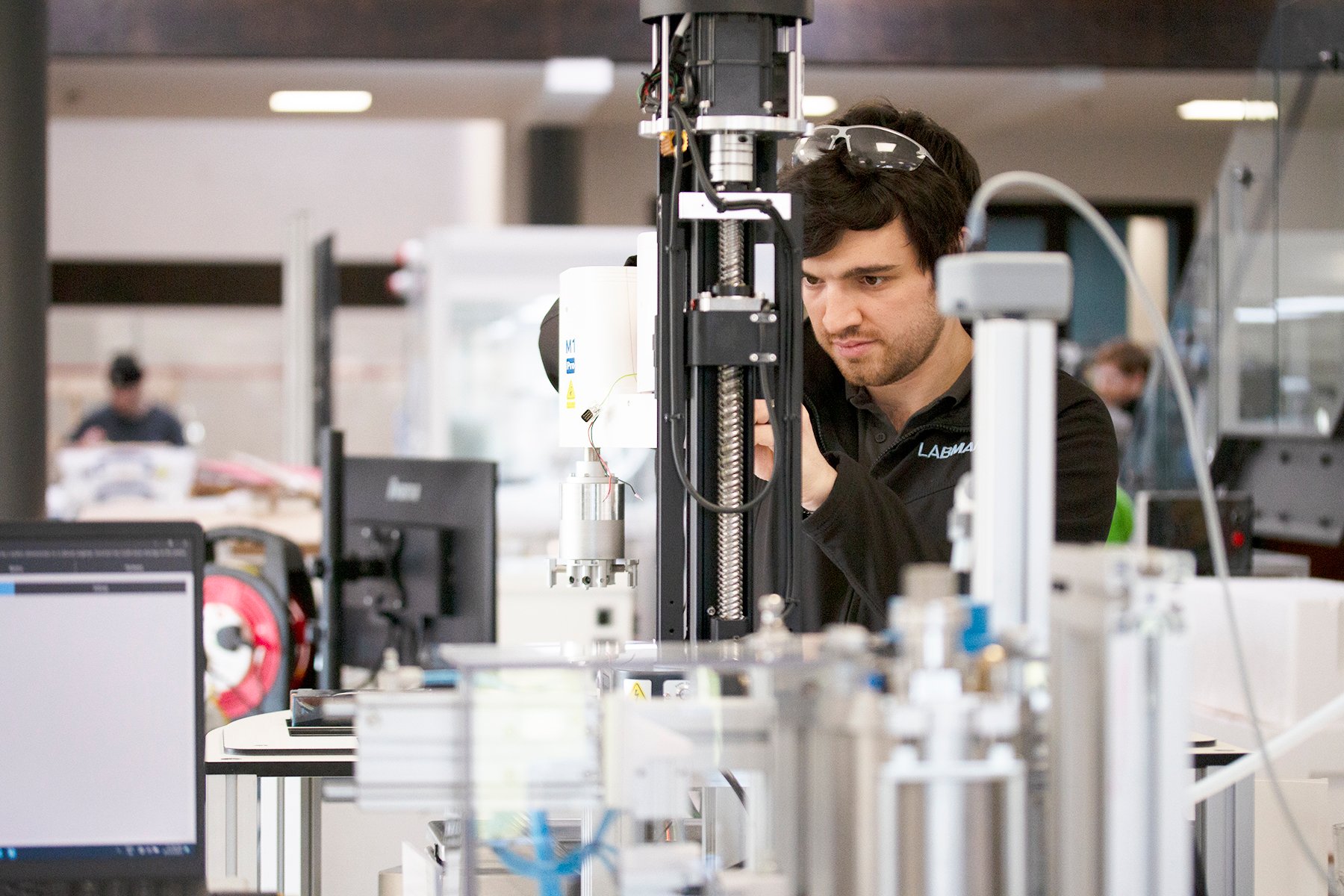 man with dark hair wearing Labman jacket and safety goggles on his head shown behind lab robotic equipment