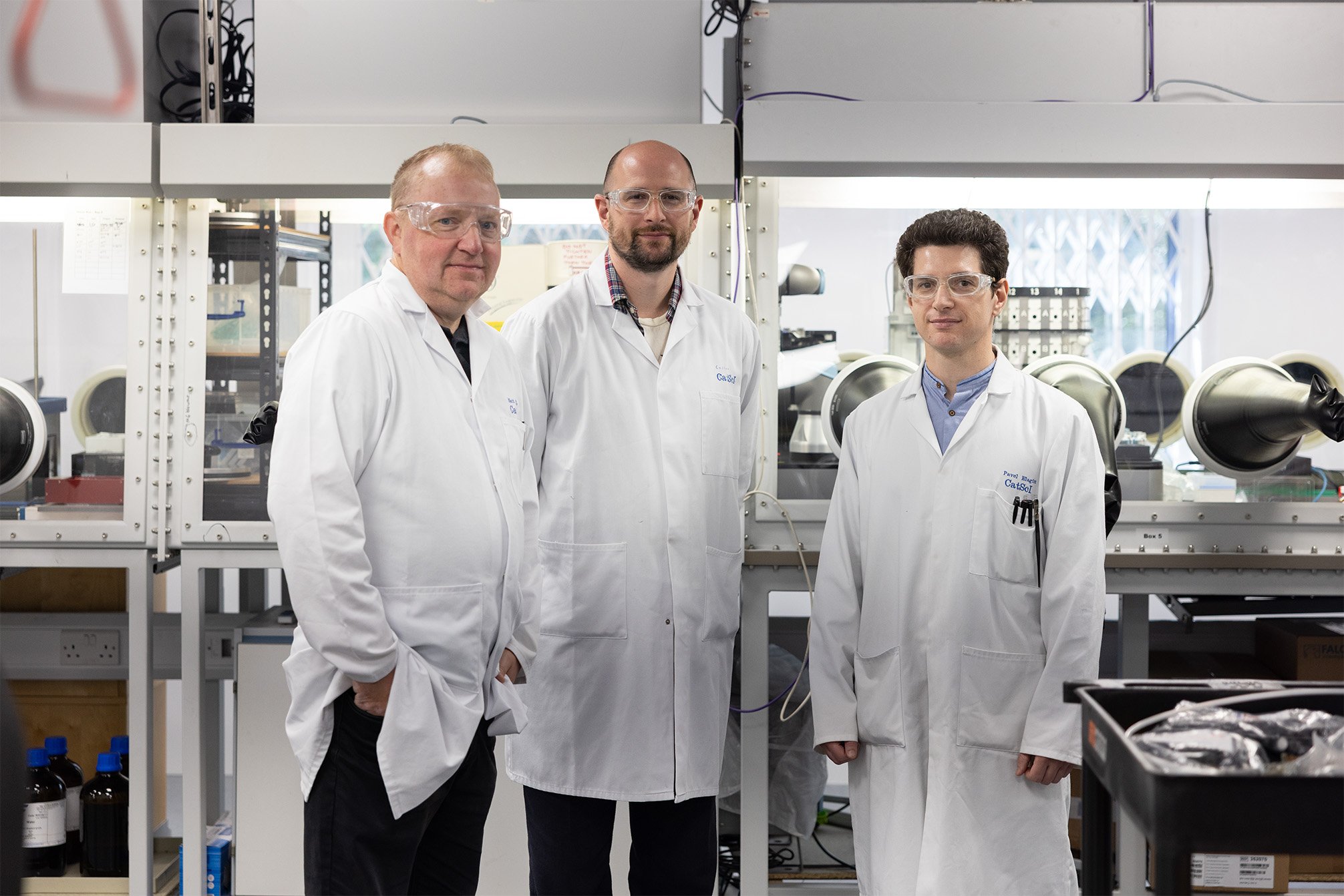 Three men wearing labcoats stood in front of a large glovebox robot in a lab