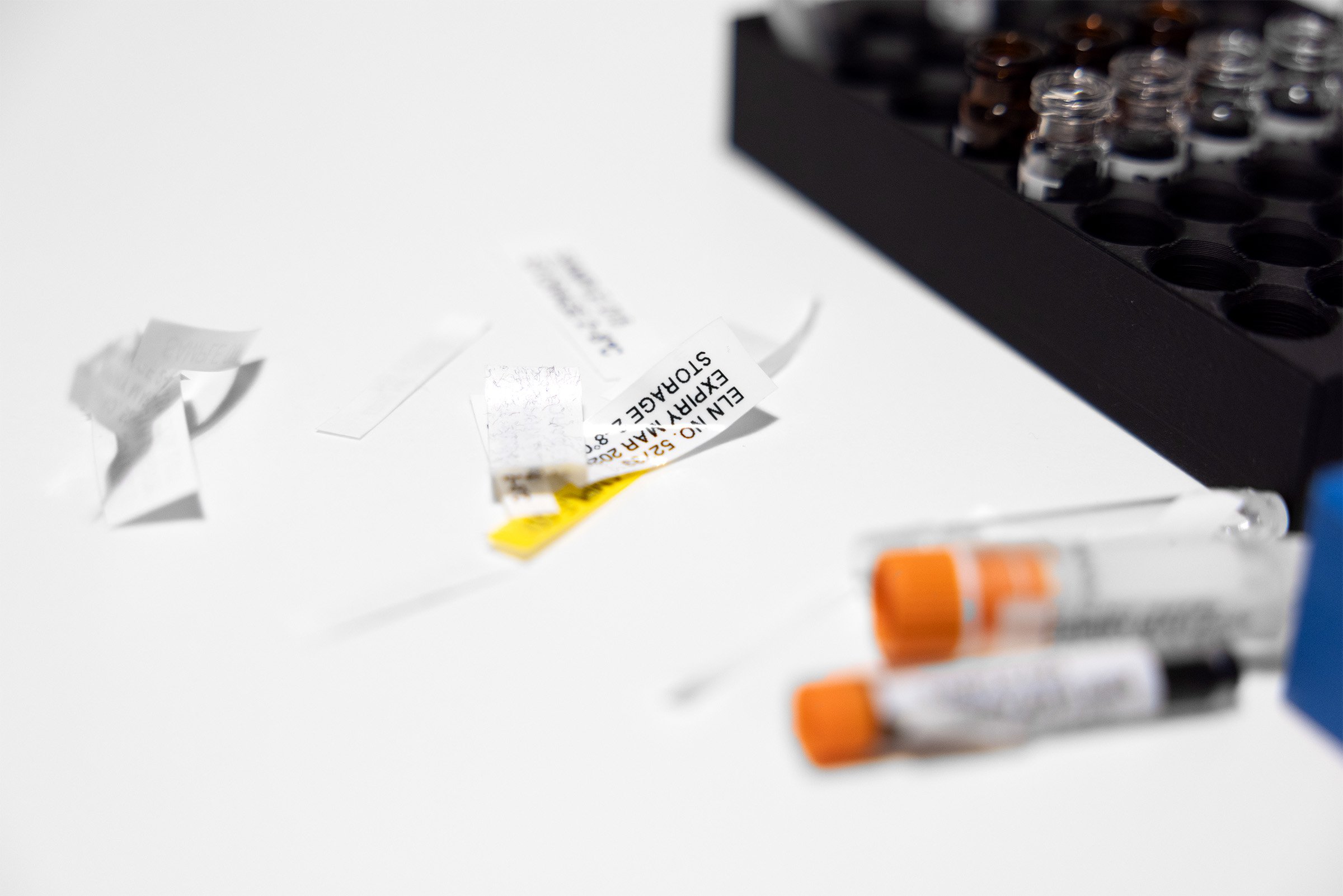 A small pile of laboratory labels and rubbish on a desk, next to a rack of vials