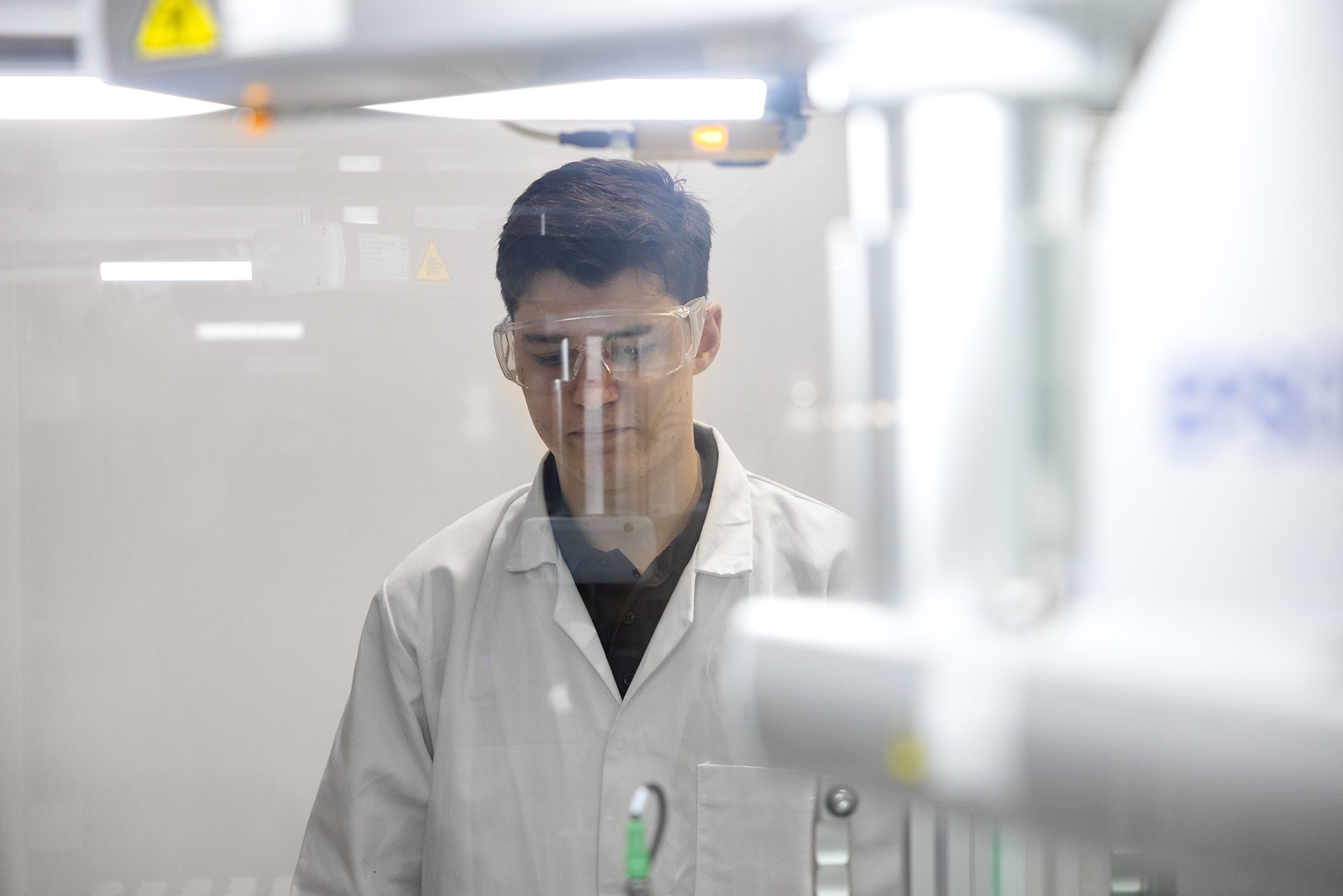 A male lab technician wearing safety specs looking through glass