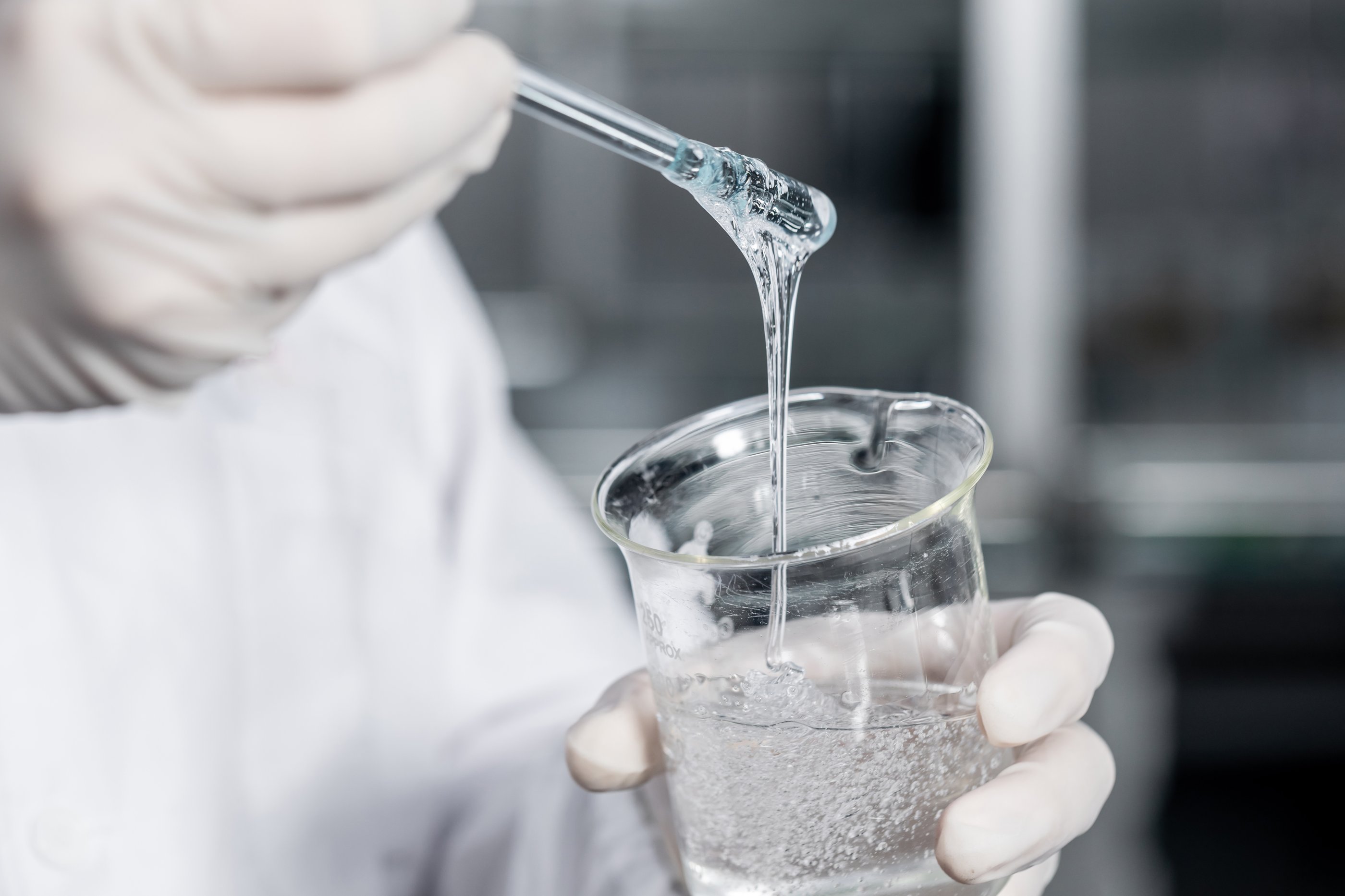 Person wearing white lab coat and white latex gloves is holding a beaker in their left containing a viscous clear glue substance. They are using a stirrer and lifting it out, the way the substance drips into the beaker suggests it is quite a thick liquid.