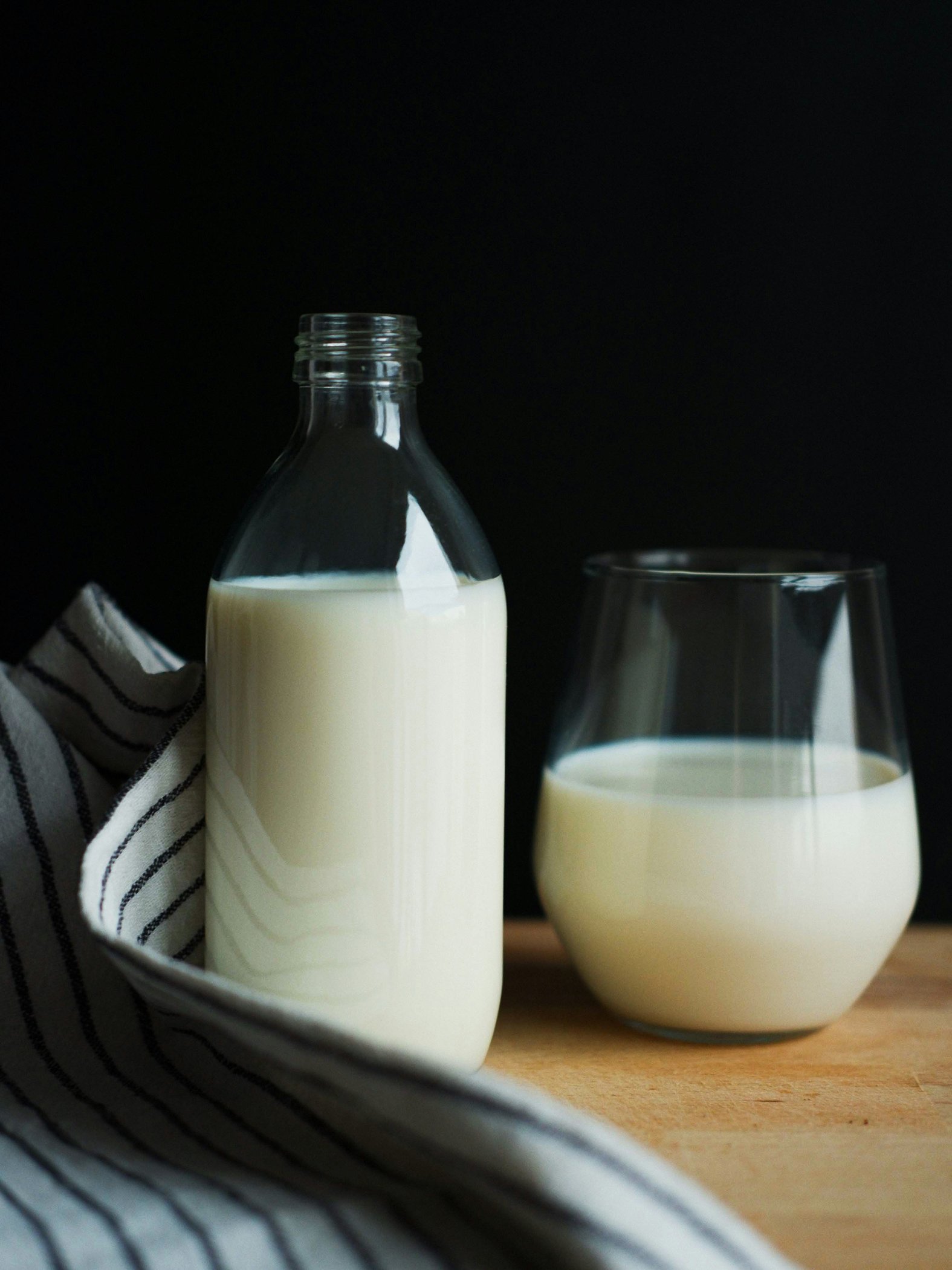 a glass bottle full of milk next to a glass full of milk sitting on a wooden table next to a linen cloth