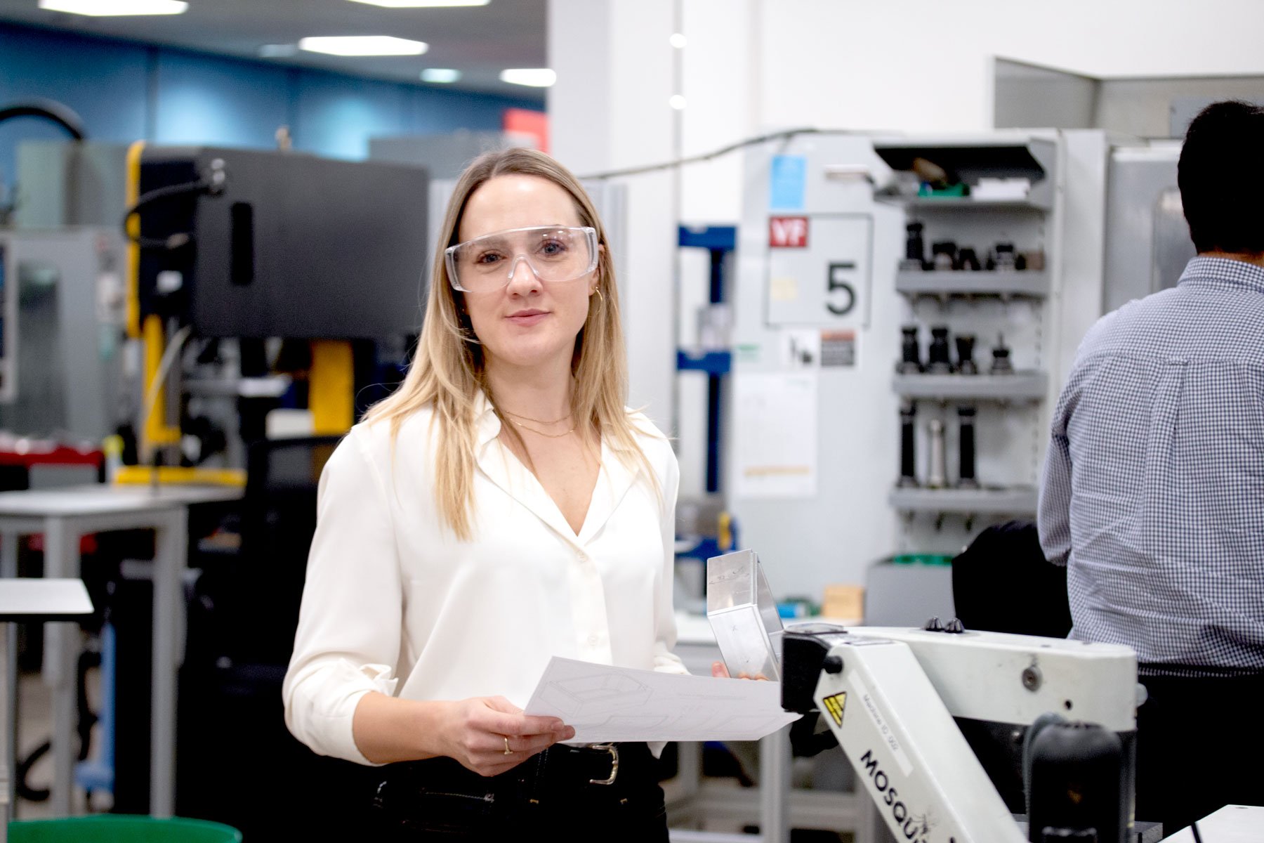 A woman in safety goggles holding up a piece of paper in front of a lab and robotic systems