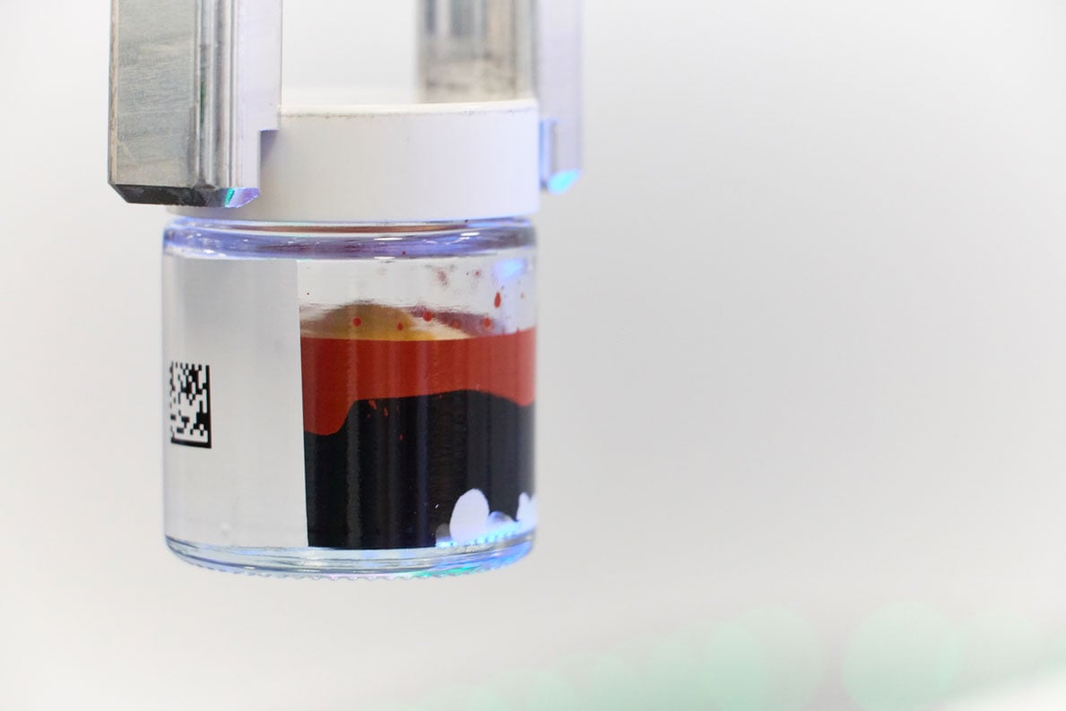 A clear pot held in a robotic gripper containing coloured, layered viscous liquid