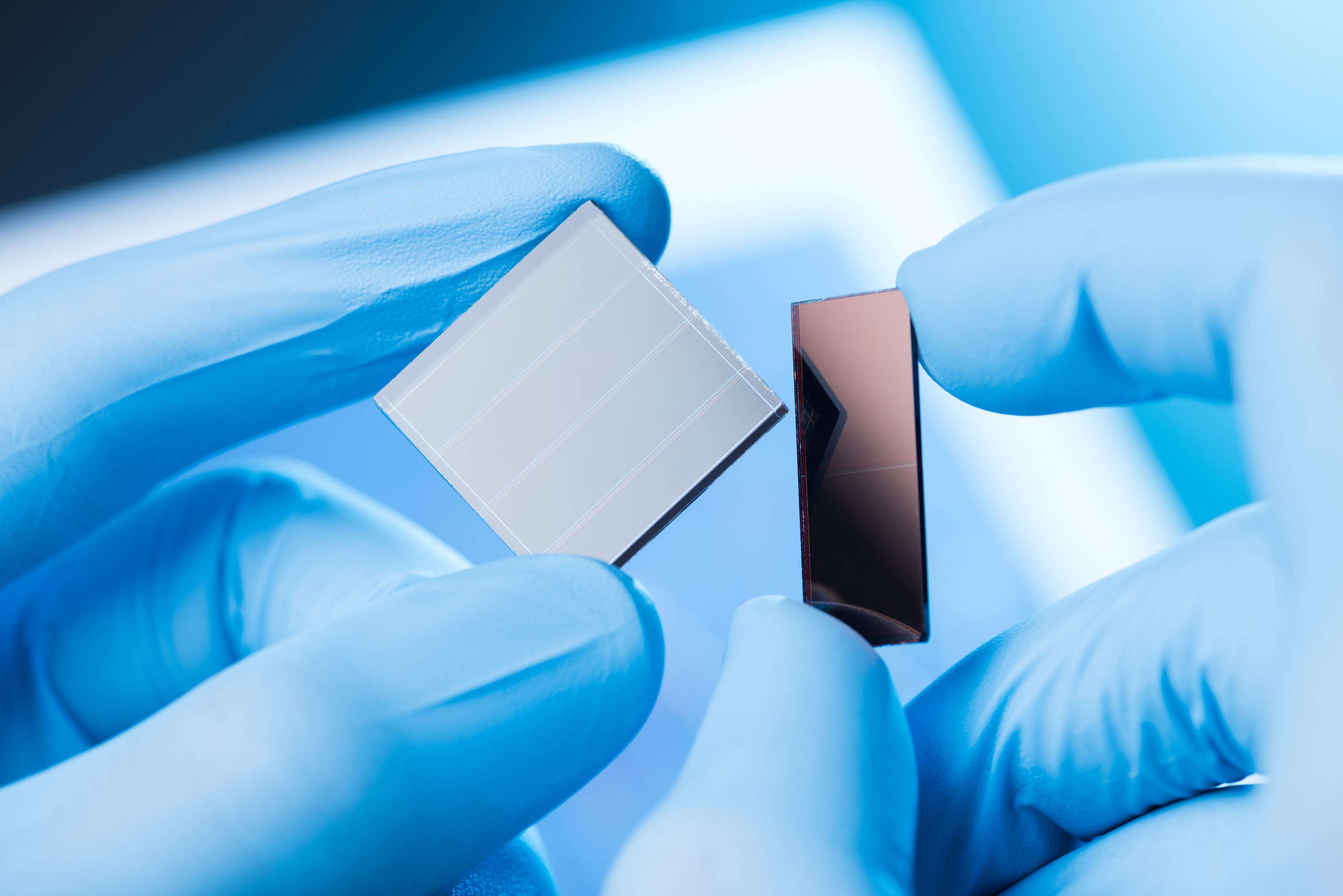 Close-up of someone wearing blue latex lab gloves holding different solar panel substrate samples in each hand.