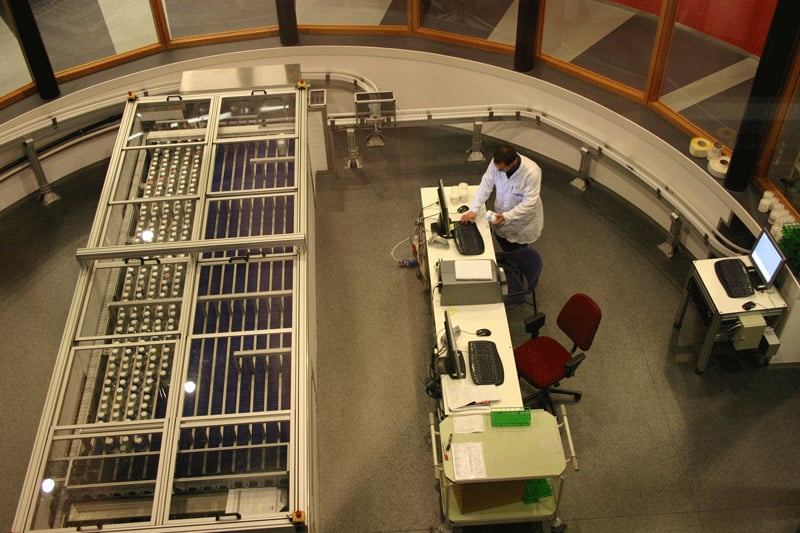Birds eye view of a robotic sample system in a round room with a long desk opposite with two laptops and a man standing at one