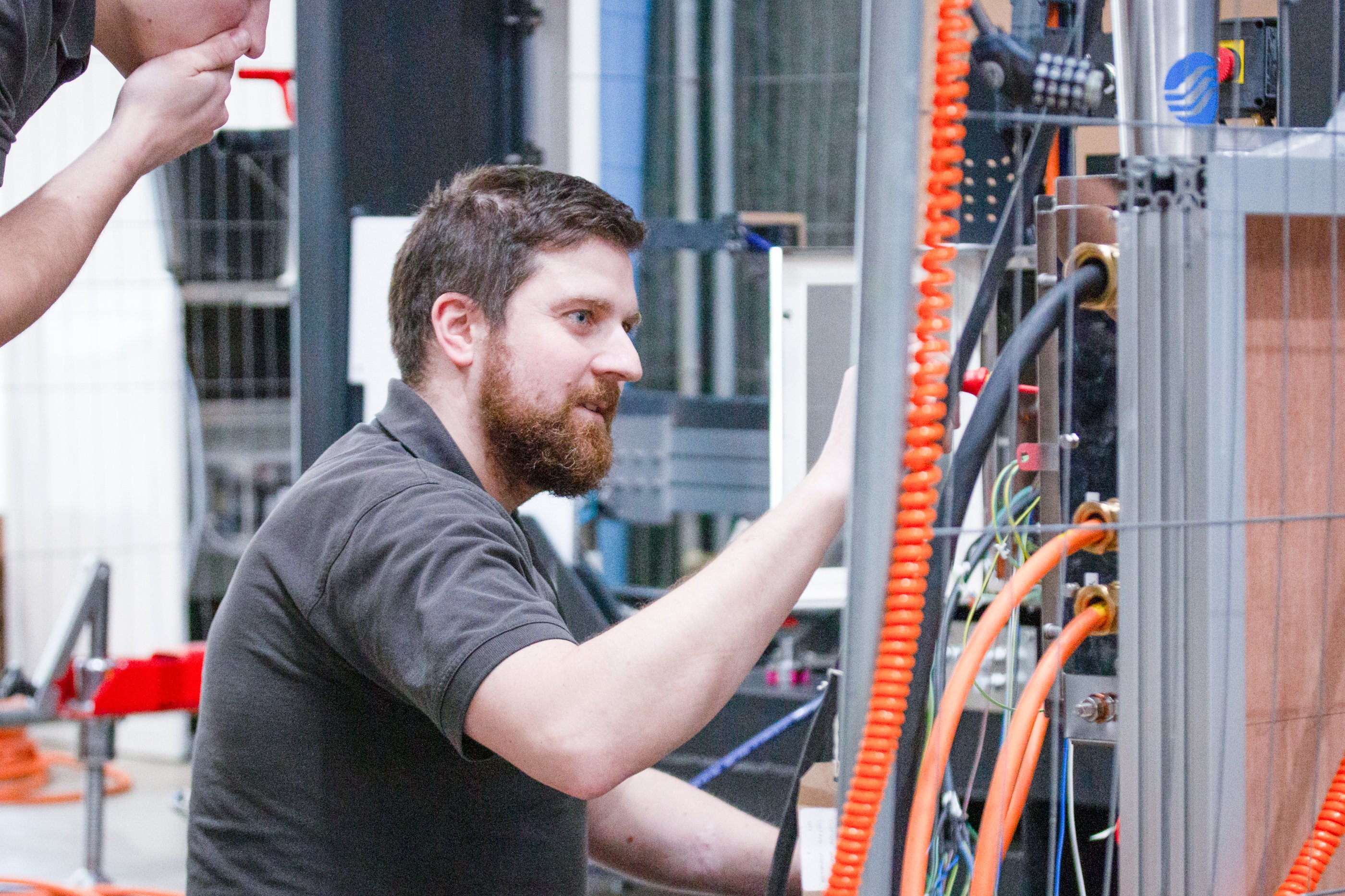 A man in a grey top in a lab looking at a machine with wires and cables coming out of it with a man standing behind with a hand over his mouth 