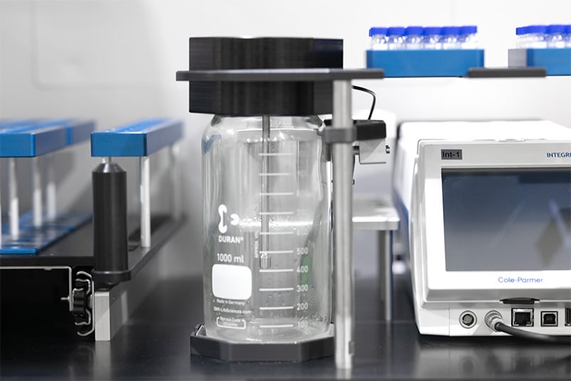 A large clear vial with a large black lid on a black table, surrounded by a blank screen and blue lidded smaller vials