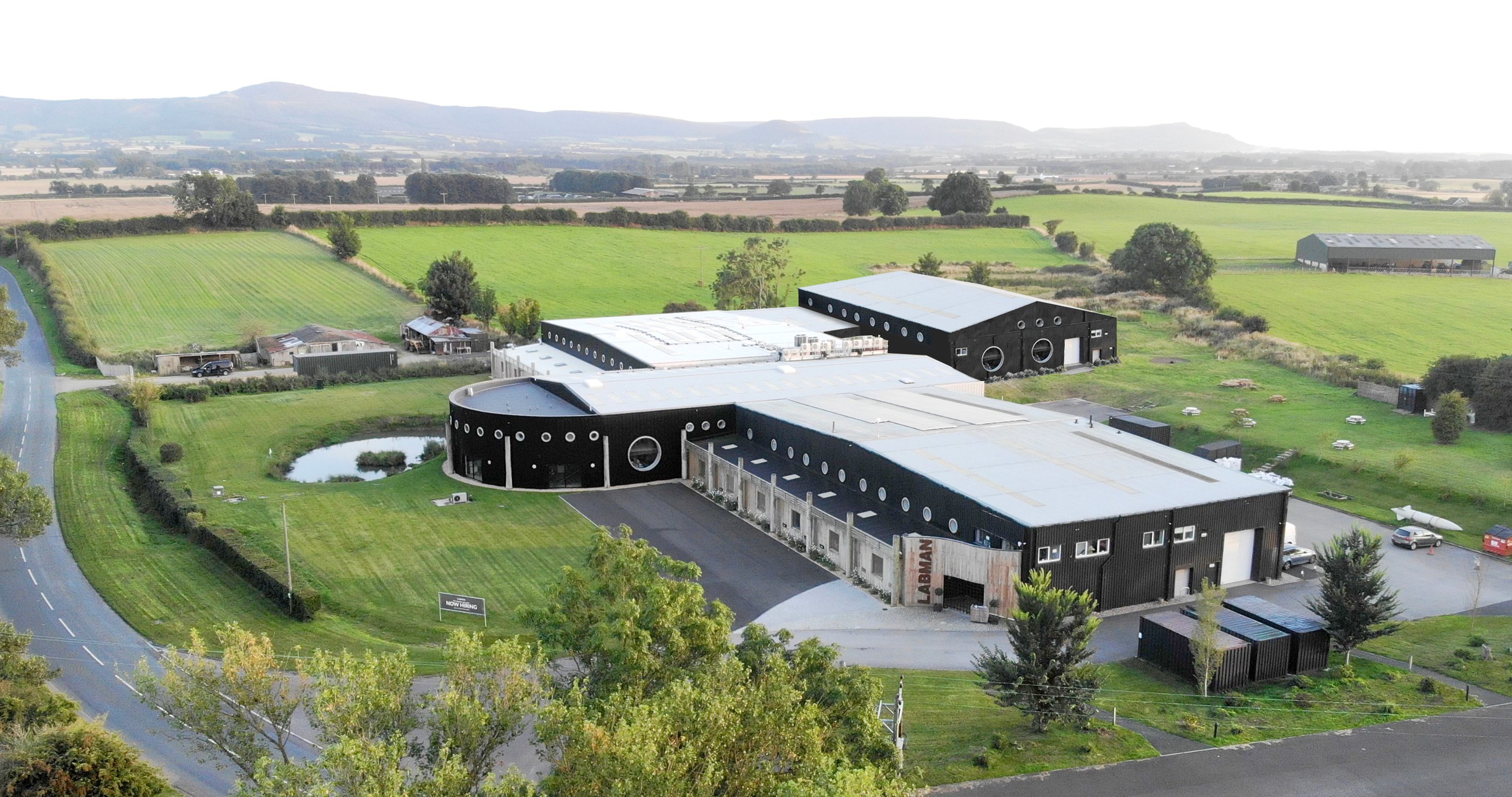A large black building with round windows in the countryside, surrounded by fields and a road to the left