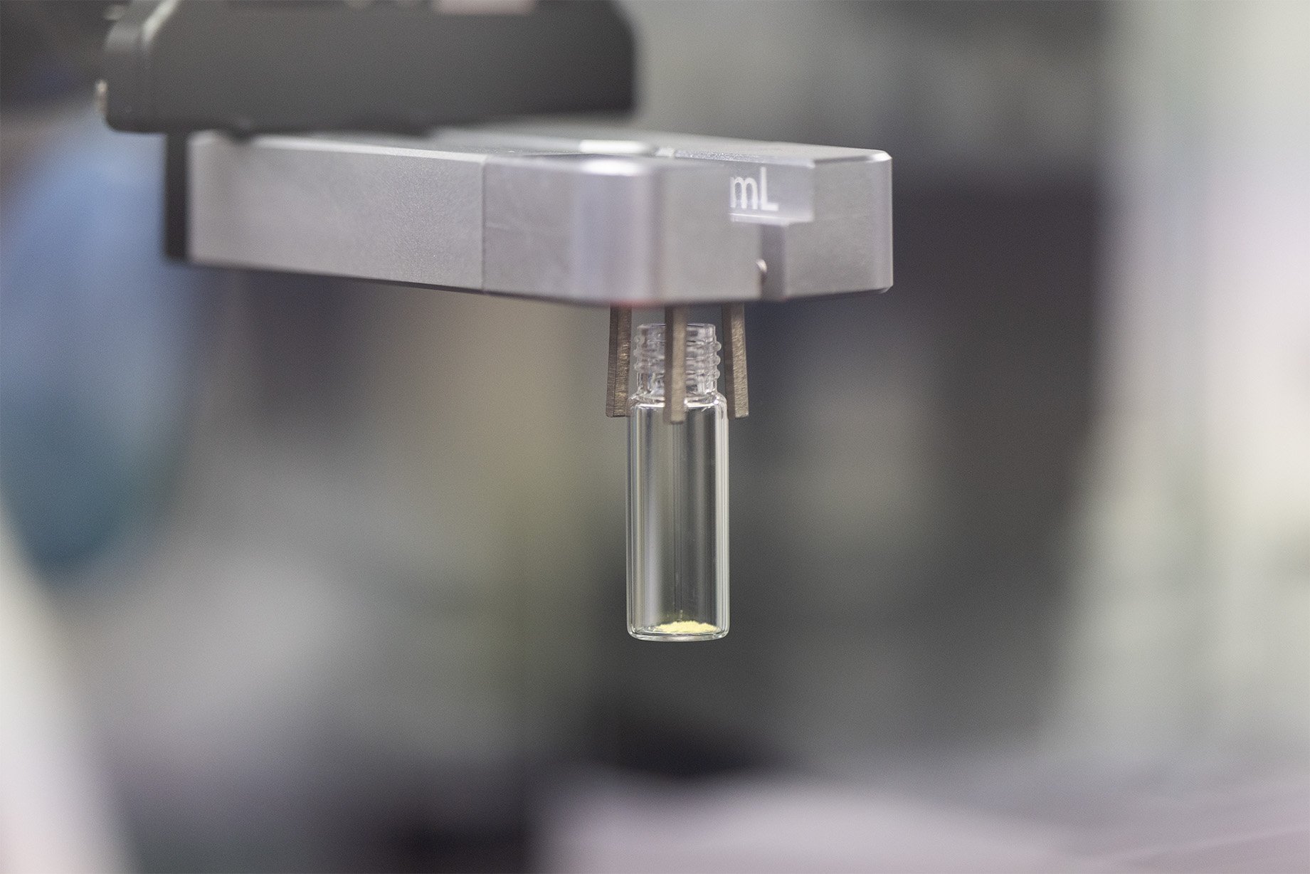 A robotic gripper holding a clear glass vial containing a small amount of powder