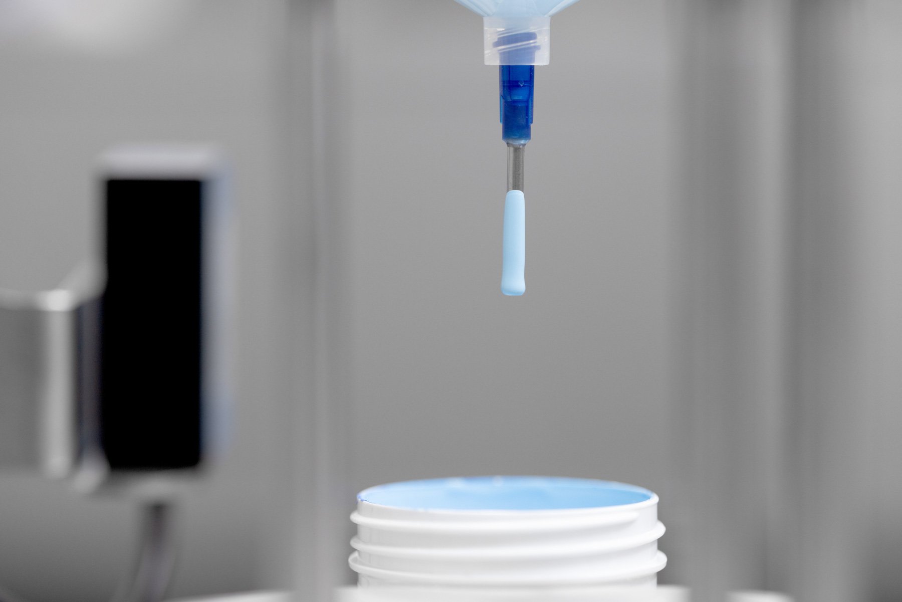 A close up of a syringe dipped in light blue paint, in a lab above a pot of the liquid
