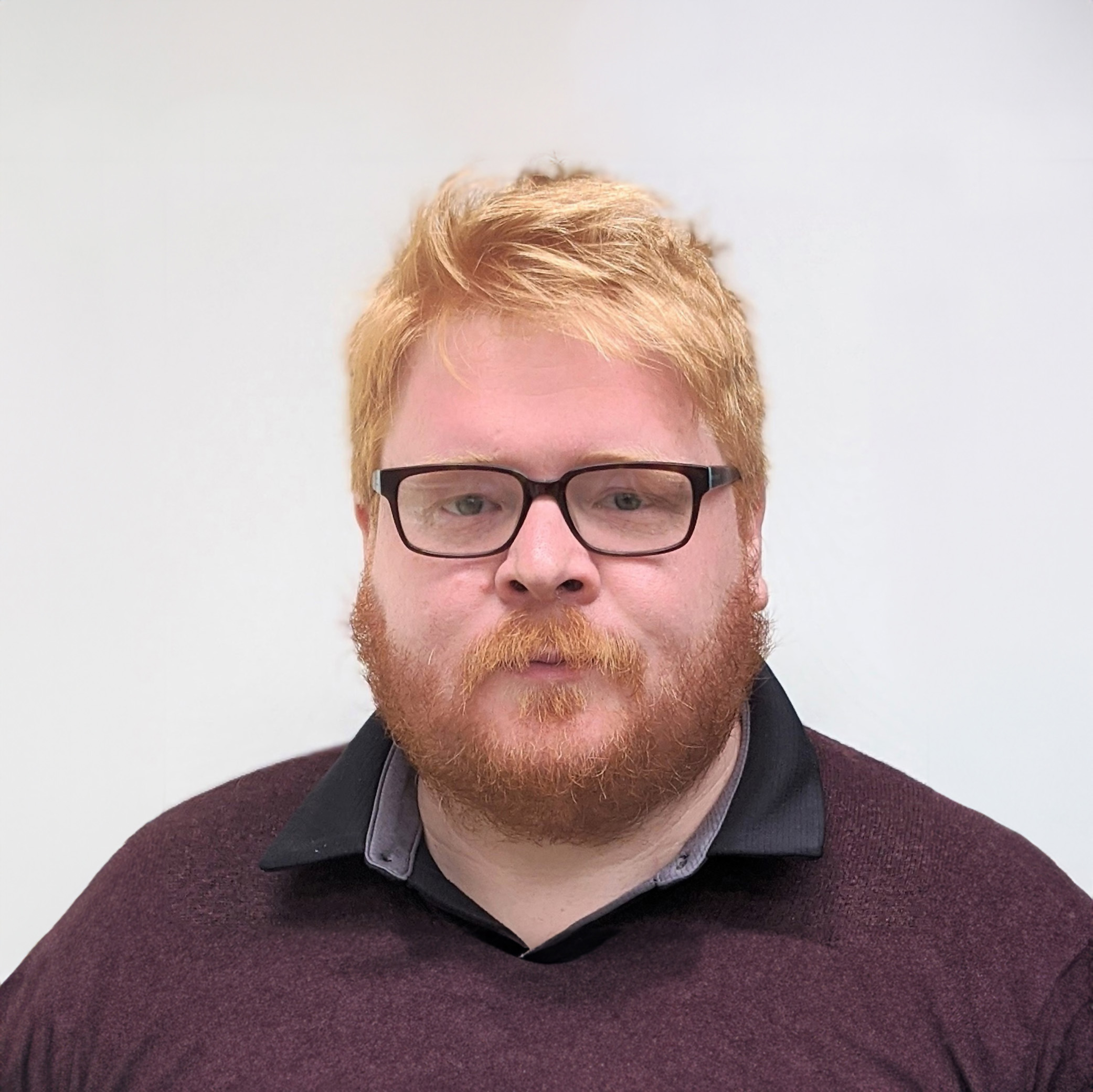 A headshot of a ginger man with glasses and a beard on a white background