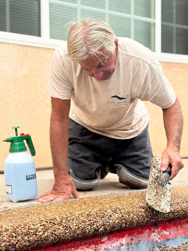 A Stone Carpet Pro's technician installing a beautiful pool deck, kneeling and using a trowel to spread the stone carpet material. A spray bottle sits beside him.