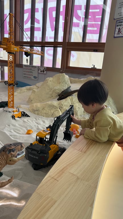 두돌 아기가 모래밭 가장자리에 앉아 굴착기 장난감을 만지며 노는 모습