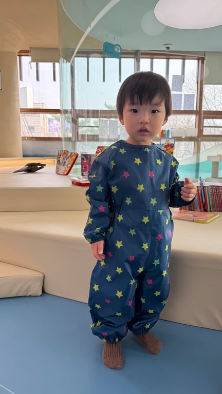 두돌 아기가 별무늬 미술가운(올인원)을 입고 서 있는 모습 — 무료 제공 가운으로 옷 걱정 없이 놀이