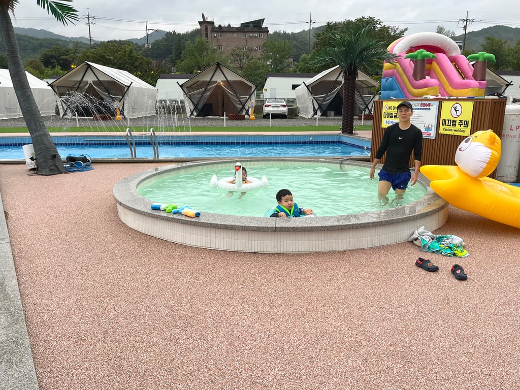포천 더세븐글램핑카라반 온수풀에서 물놀이하는 아이와 아빠 — 뒤로 에어바운스와 글램핑 텐트가 보인다