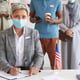 Multi-ethnic group of people standing in row and wearing masks at polling station on election day, focus on modern senior woman  while registering for voting, copy space