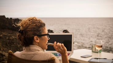 Rear view of woman looking away while sitting with laptop and alcohol at beach during sunset