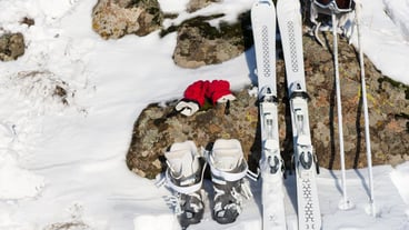 Winter sports setup displaying red gloves, pair of skis, boots, helmet and poles leaning on surface of stone partially covered with snow