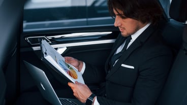 Using notepad. Young businessman in black suit and tie inside modern automobile.