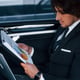 Using notepad. Young businessman in black suit and tie inside modern automobile.