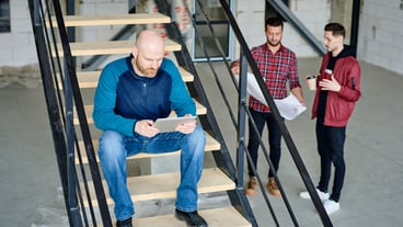 Bald engineer with digital tablet sitting on staircase and scrolling through online sketch while his colleagues discussing blueprint
