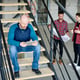 Bald engineer with digital tablet sitting on staircase and scrolling through online sketch while his colleagues discussing blueprint